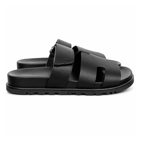 Side profile of the Hermès Chypre sandal crafted in smooth black calfskin leather, featuring the signature 'H' cut-out design across the toe, an adjustable velcro strap over the instep, and a contoured anatomical rubber sole.