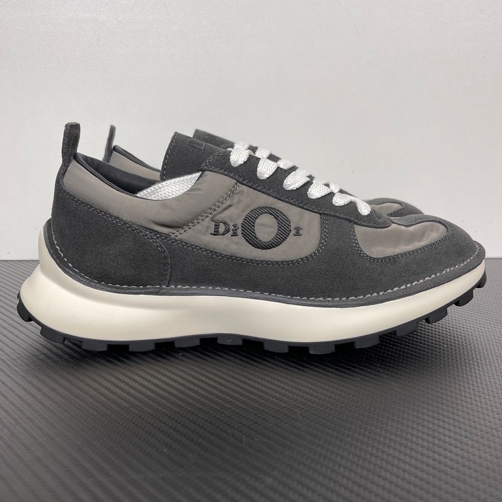 Side profile of the Dior Squash sneaker in a black and grey colorway, featuring a mix of premium calf suede and technical nylon, distinctive 'Dior' side embroidery with an oversized 'O', and a chunky white midsole with a lugged black outsole.