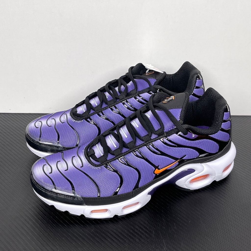 Side profile of the Nike Air Max Plus OG sneaker featuring a vibrant Voltage Purple gradient mesh upper, signature black wavy TPU overlays, striking Total Orange accents on the Swoosh and visible Air units, resting on a crisp white midsole.