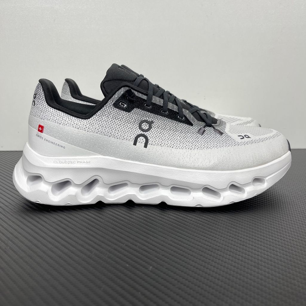 Side profile of the On Running Cloudtilt sneaker featuring a sleek, lightweight black knit upper with a sock-like collar, integrated speed lacing, and a contrasting ivory midsole showcasing the collapsing CloudTec Phase® pods.