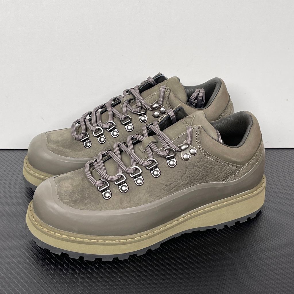 Side profile of the UMMCOR low-top hiking shoe in an earthy Army Green colorway, featuring a textured cowhide leather upper, a protective wrap-around rubber mudguard, metal D-ring lacing hardware, and a chunky lugged rubber outsole.