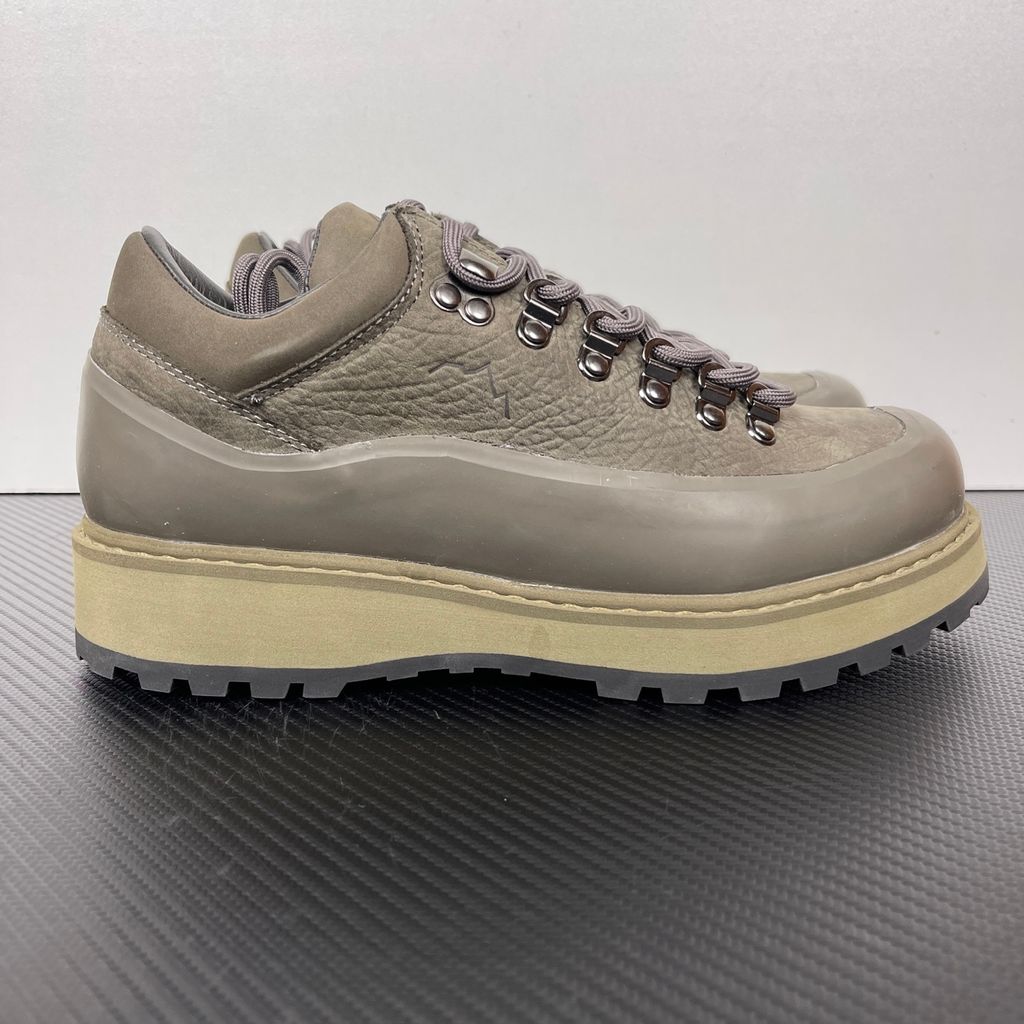 Side profile of the UMMCOR low-top hiking shoe in an earthy Army Green colorway, featuring a textured cowhide leather upper, a protective wrap-around rubber mudguard, metal D-ring lacing hardware, and a chunky lugged rubber outsole.