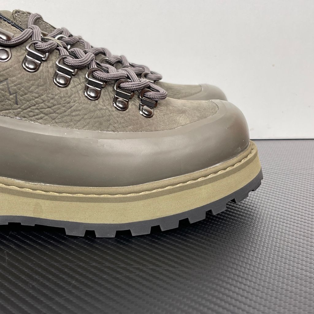 Side profile of the UMMCOR low-top hiking shoe in an earthy Army Green colorway, featuring a textured cowhide leather upper, a protective wrap-around rubber mudguard, metal D-ring lacing hardware, and a chunky lugged rubber outsole.