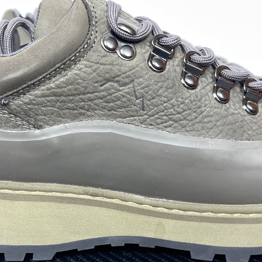 Side profile of the UMMCOR low-top hiking shoe in an earthy Army Green colorway, featuring a textured cowhide leather upper, a protective wrap-around rubber mudguard, metal D-ring lacing hardware, and a chunky lugged rubber outsole.