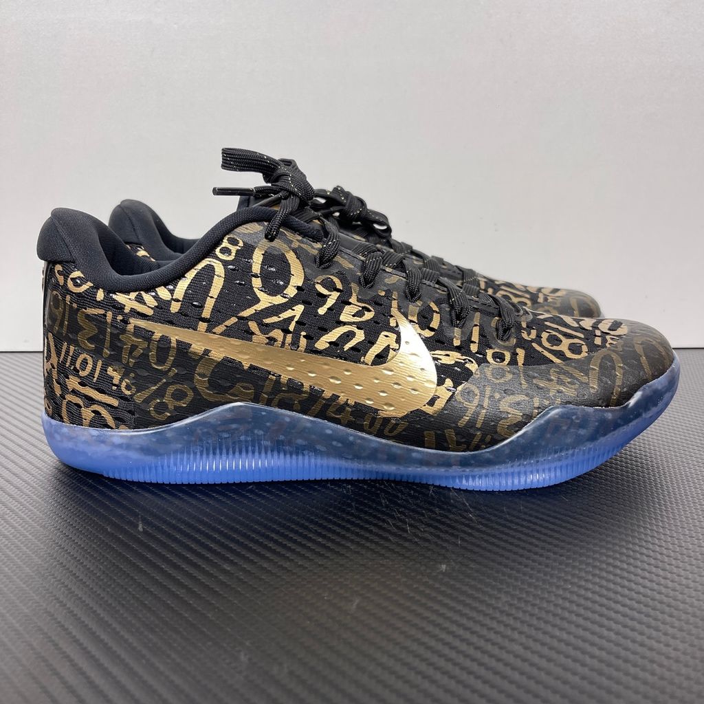 Side profile of the Nike Kobe 11 Protro basketball shoe in the Mamba Day colorway, featuring a sleek black upper with striking metallic gold Swoosh detailing, signature Kobe branding, and a translucent rubber outsole.