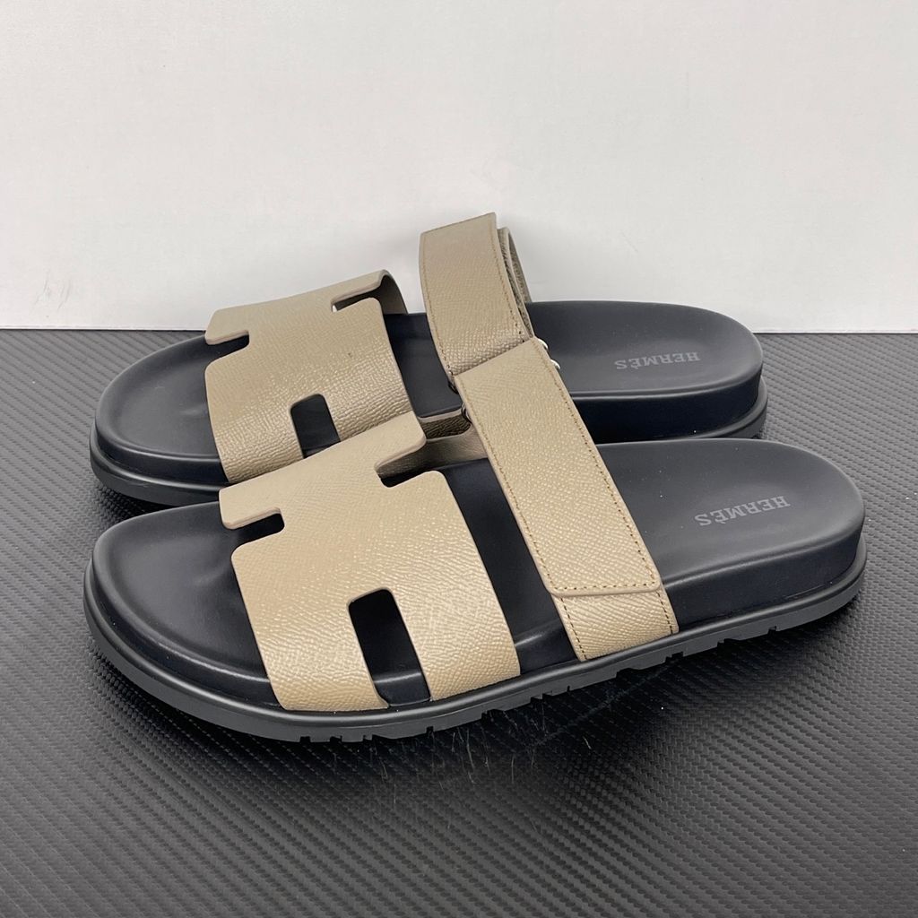 Side profile of the Hermès Chypre Sandal featuring a textured taupe leather upper with the signature 'H' cutout, an adjustable velcro strap, and a comfortable, anatomically molded black rubber footbed.