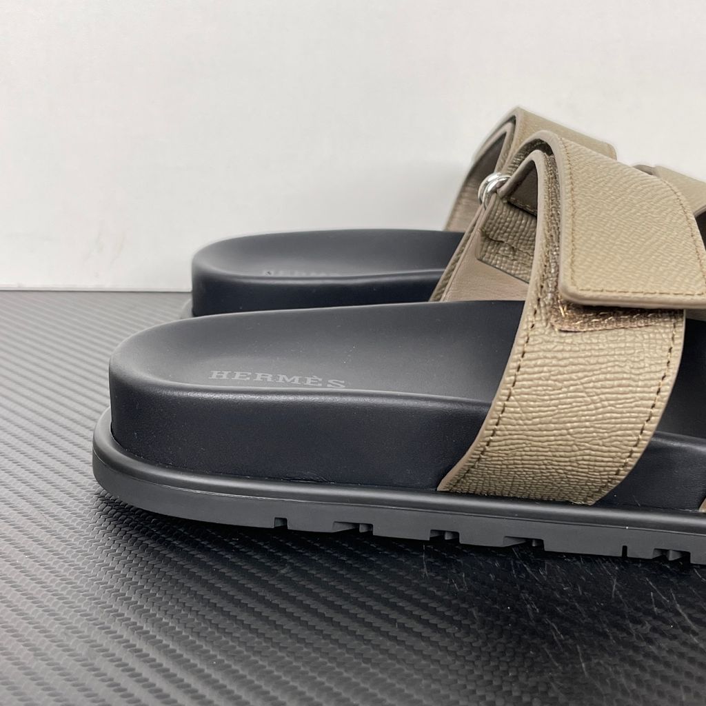 Side profile of the Hermès Chypre Sandal featuring a textured taupe leather upper with the signature 'H' cutout, an adjustable velcro strap, and a comfortable, anatomically molded black rubber footbed.