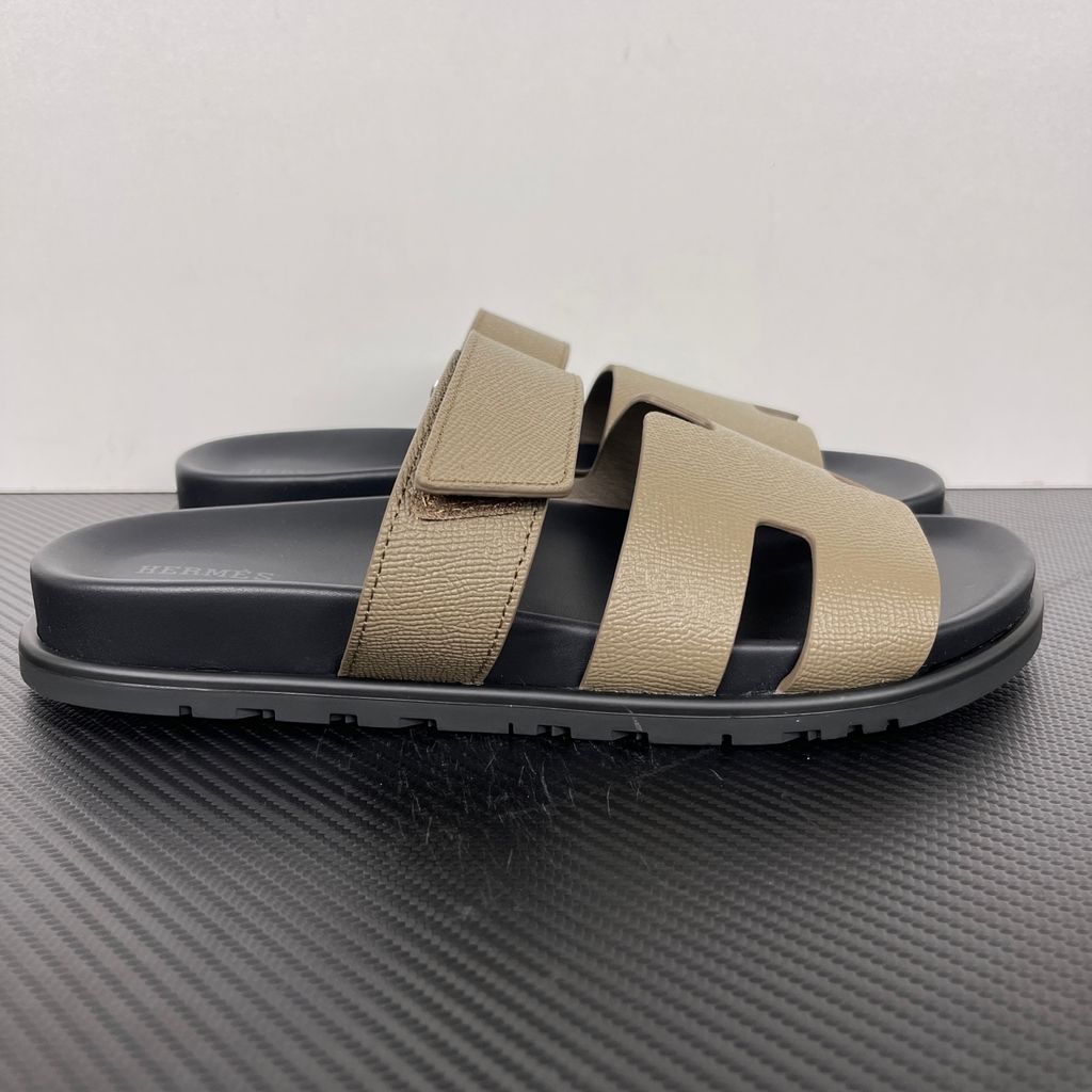 Side profile of the Hermès Chypre Sandal featuring a textured taupe leather upper with the signature 'H' cutout, an adjustable velcro strap, and a comfortable, anatomically molded black rubber footbed.
