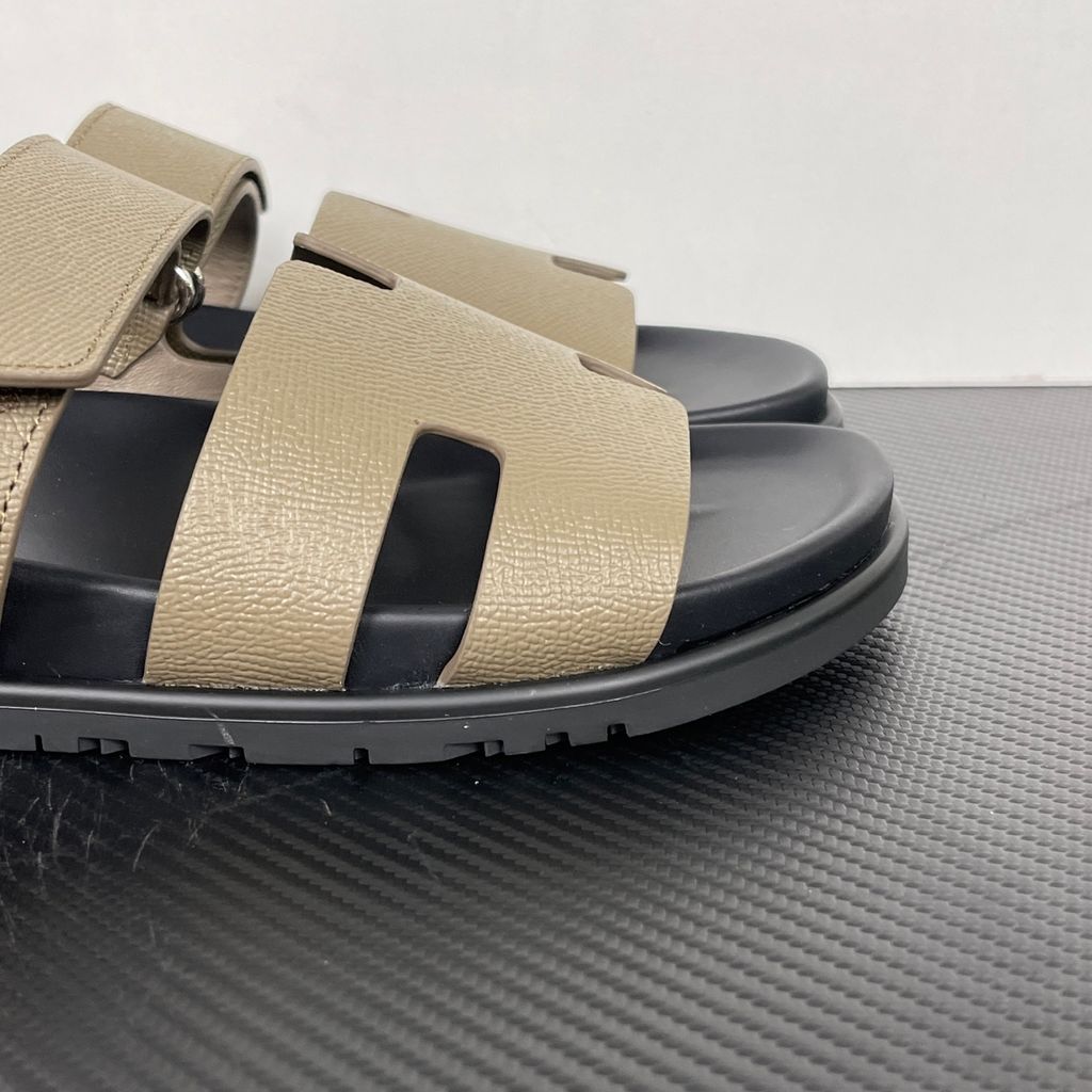 Side profile of the Hermès Chypre Sandal featuring a textured taupe leather upper with the signature 'H' cutout, an adjustable velcro strap, and a comfortable, anatomically molded black rubber footbed.