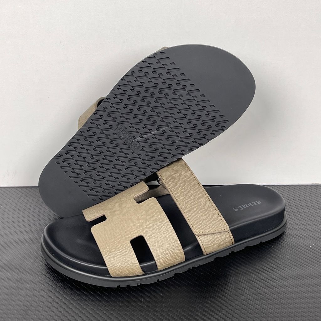 Side profile of the Hermès Chypre Sandal featuring a textured taupe leather upper with the signature 'H' cutout, an adjustable velcro strap, and a comfortable, anatomically molded black rubber footbed.