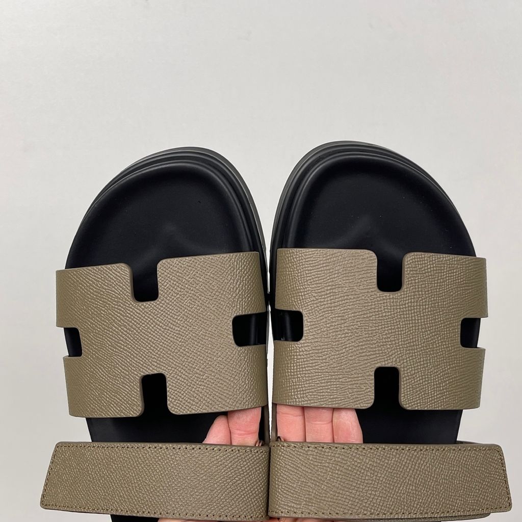 Side profile of the Hermès Chypre Sandal featuring a textured taupe leather upper with the signature 'H' cutout, an adjustable velcro strap, and a comfortable, anatomically molded black rubber footbed.