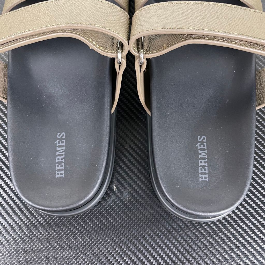 Side profile of the Hermès Chypre Sandal featuring a textured taupe leather upper with the signature 'H' cutout, an adjustable velcro strap, and a comfortable, anatomically molded black rubber footbed.