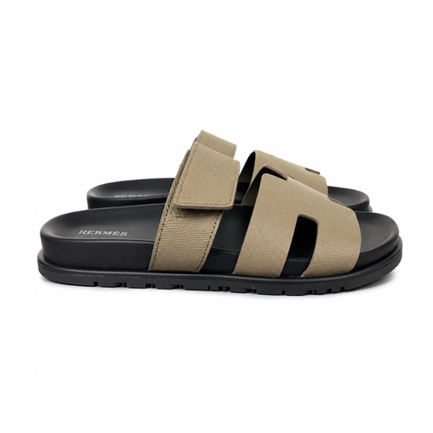 Side profile of the Hermès Chypre Sandal featuring a textured taupe leather upper with the signature 'H' cutout, an adjustable velcro strap, and a comfortable, anatomically molded black rubber footbed.