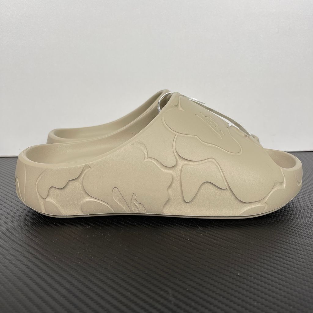 Side profile of the beige A Bathing Ape (BAPE) foam slide (image_889658.jpg), featuring a chunky molded silhouette, a smooth slip-on upper, and deeply debossed signature ape camo patterns wrapping around the midsole and foot strap.