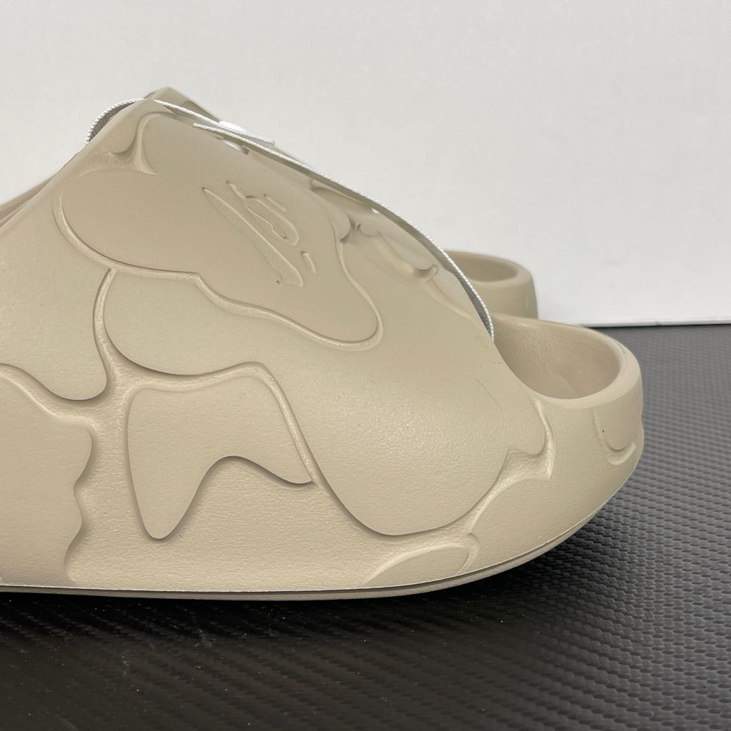 Side profile of the beige A Bathing Ape (BAPE) foam slide (image_889658.jpg), featuring a chunky molded silhouette, a smooth slip-on upper, and deeply debossed signature ape camo patterns wrapping around the midsole and foot strap.