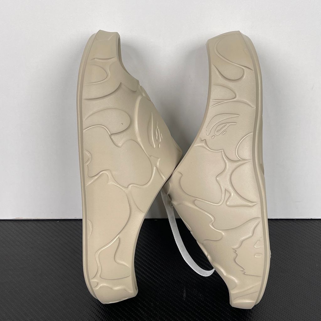 Side profile of the beige A Bathing Ape (BAPE) foam slide (image_889658.jpg), featuring a chunky molded silhouette, a smooth slip-on upper, and deeply debossed signature ape camo patterns wrapping around the midsole and foot strap.