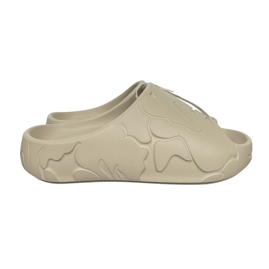 Side profile of the beige A Bathing Ape (BAPE) foam slide (image_889658.jpg), featuring a chunky molded silhouette, a smooth slip-on upper, and deeply debossed signature ape camo patterns wrapping around the midsole and foot strap.