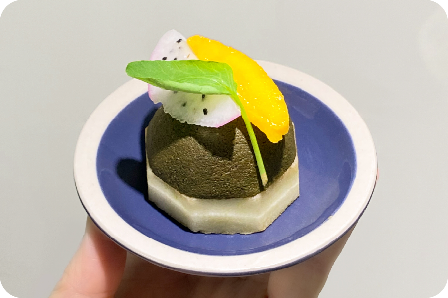 A hay powder treat shaped into a small cake and topped with fruit, on a blue plate