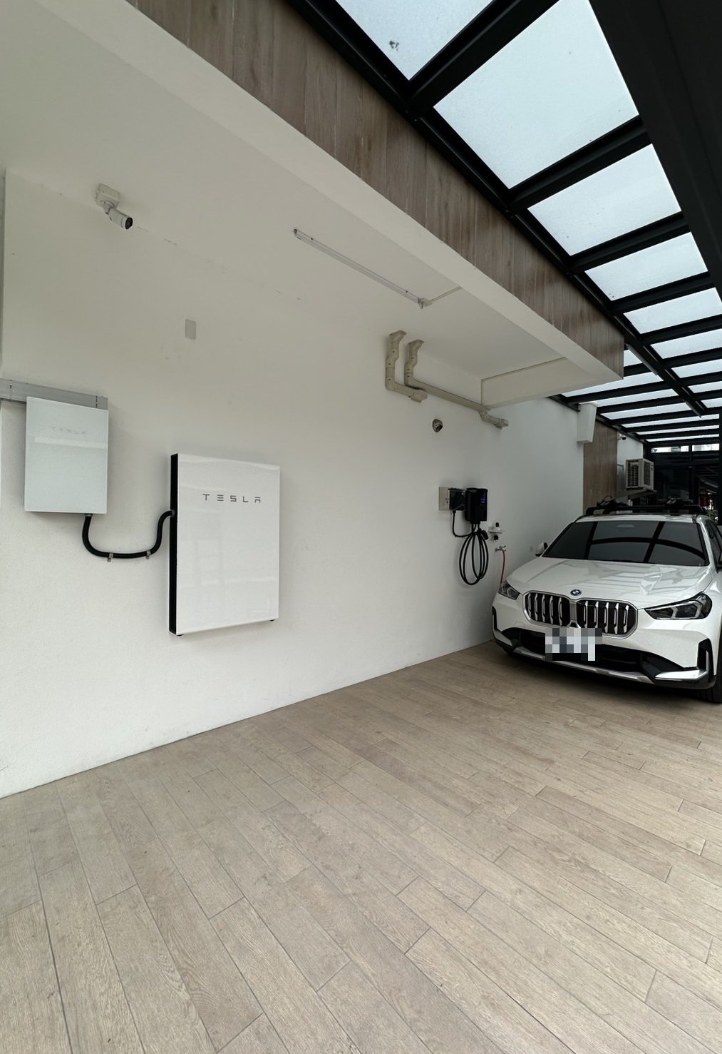 崧象智能在透天車庫安裝的 Tesla Backup Gateway（左）與 Powerwall（右），壁掛在玻璃雨遮下的牆面，右邊牆上是電動車充電座，從側面拍攝