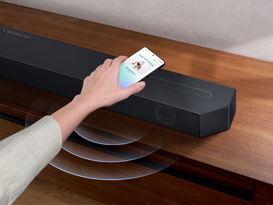 Description: A hand taps a smartphone with the Samsung music app on-screen on the Soundbar. It instantly plays music, showing how easy it is to switch from smartphone to Soundbar.