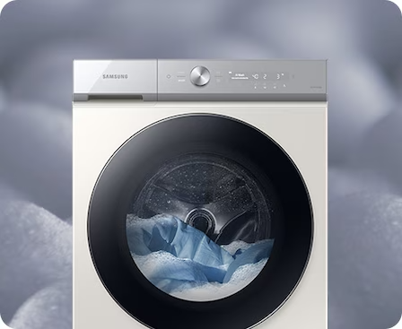 Description: A front facing white Samsung Bespoke Grande AI washer is set with detergent foam. The washer is in mid-cycle, with laundry and detergent bubbles inside the machine.