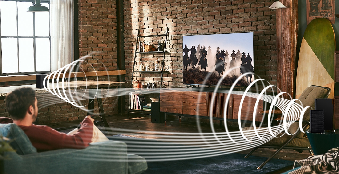 Description: A man enjoys racing content on his TV. Soundwave graphics are playing from Samsung Wireless Rear Speaker Kit and Soundbar, demonstrating Wireless Surround Sound Compatible feature of Samsung soundbar.