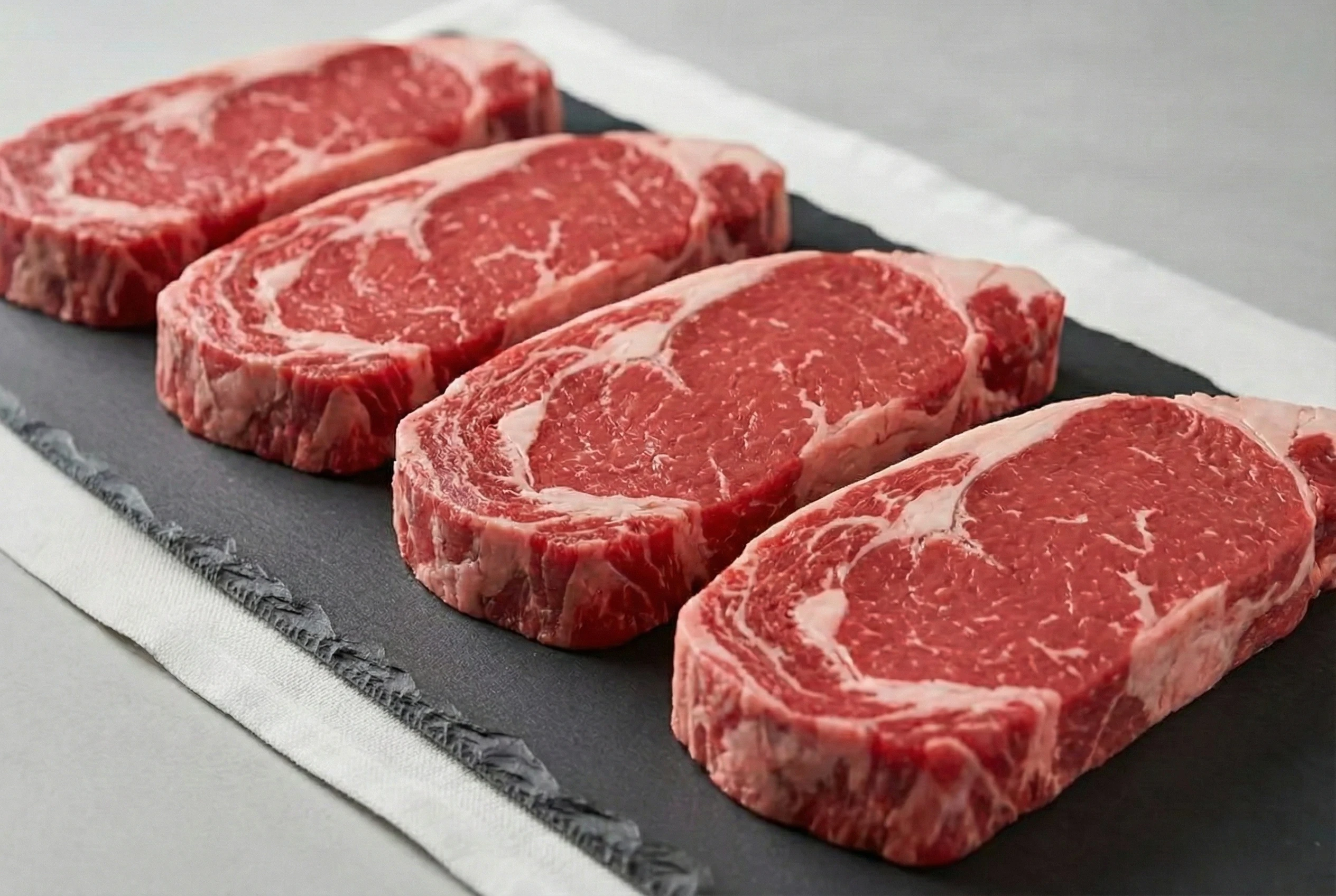 Three raw ribeye steaks from low to high intramuscular marbling on light stone.