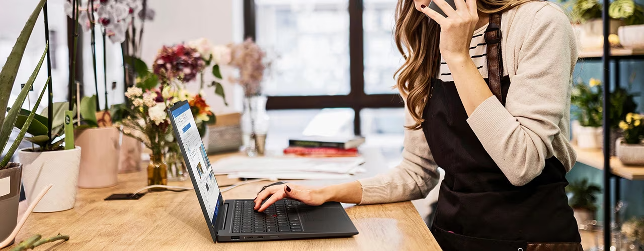 A woman is on her smartphone, typing with her right hand on the Lenovo ThinkPad X1 Carbon Gen 12 laptop, which is on a table surrounded by plants.