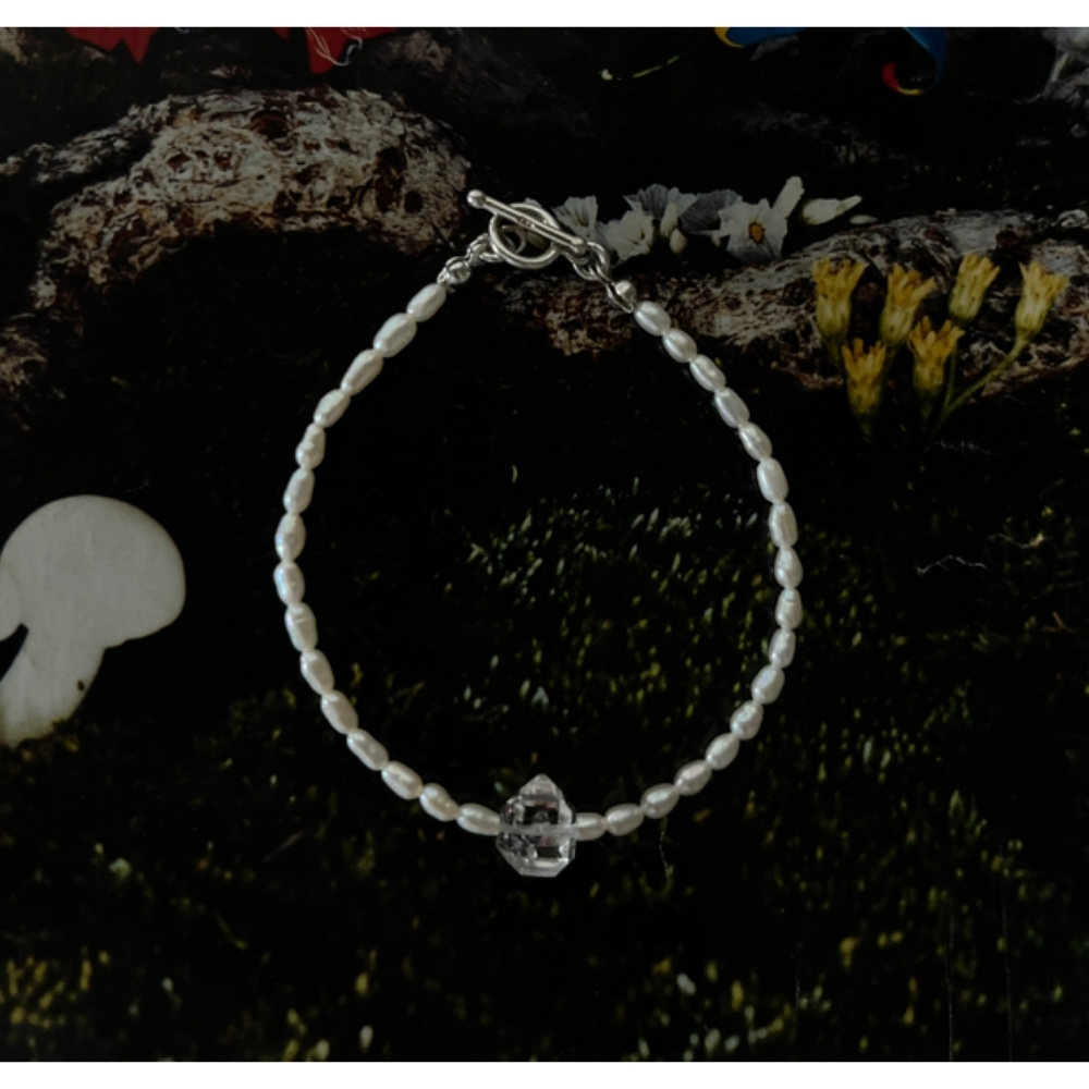 Matilda & Co.的天然米粒珍珠與赫基蒙925純銀手鍊放置在深綠色的苔蘚上，點綴著黃色小花與白色蕈菇。米粒珍珠與剔透的赫基蒙水晶在深色背景襯托下，顯得格外潔白明亮。