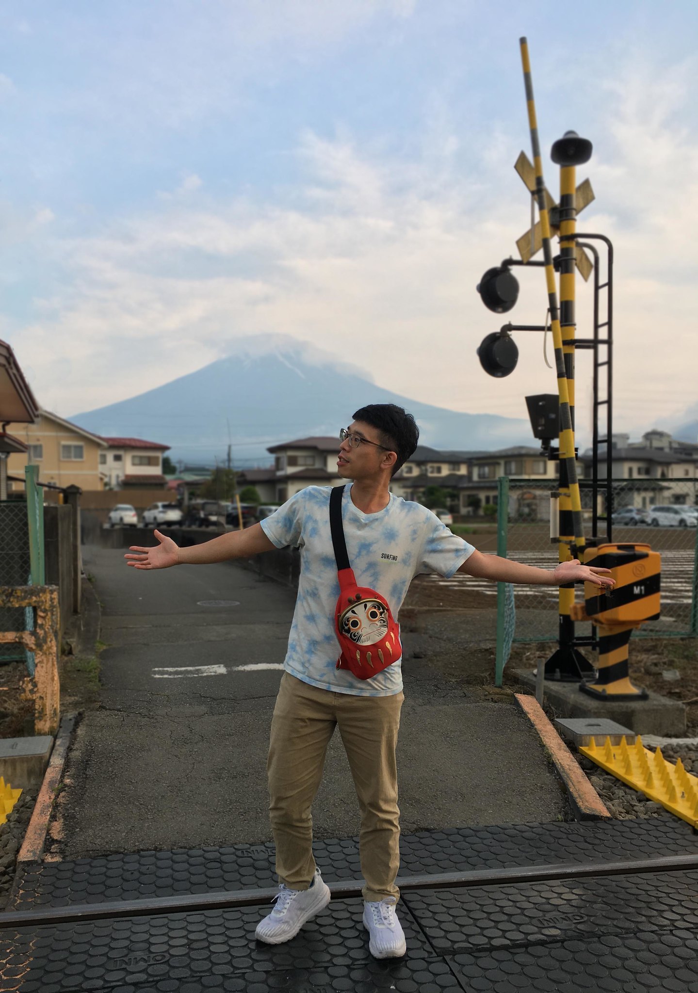 短髮男生穿著水藍色短袖上衣，揹著黑色達摩口金包，站在富士山前面的鐵道小路上
