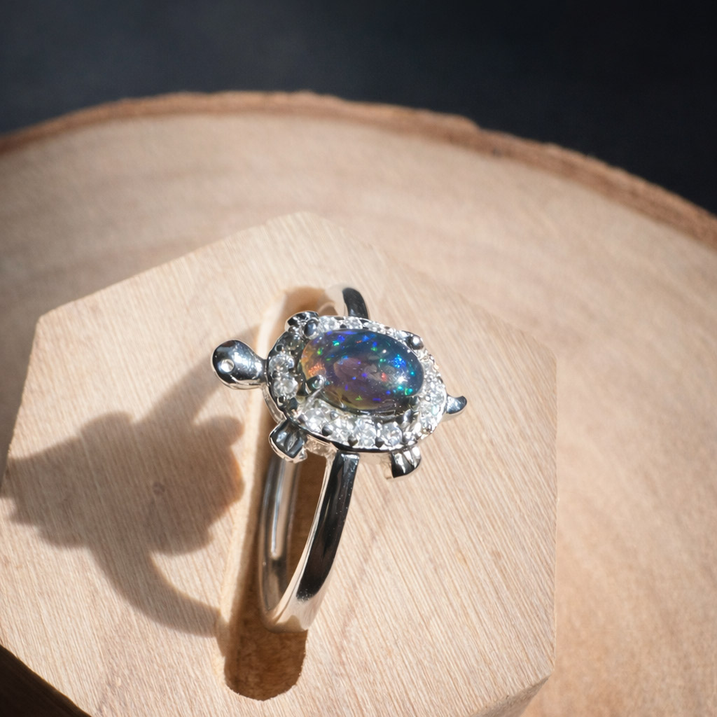 歐泊彩石海龜戒指置於木質底座上,自然光下呈現藍綠與彩虹變彩光澤,銀色海龜造型搭配細緻鑲鑽,散發沉穩療癒氣息