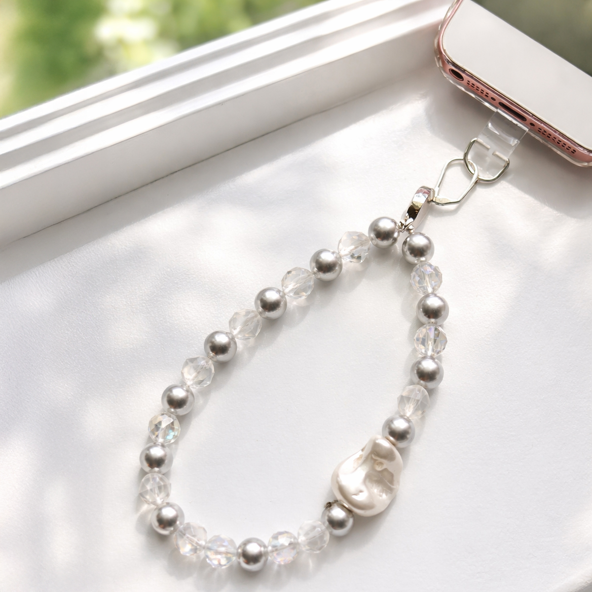 窗邊自然光下的清透白水晶與灰珍珠手機掛繩,透明水晶切面珠與灰珍珠交錯設計,搭配不規則白色珍珠墜飾,風格簡約清新