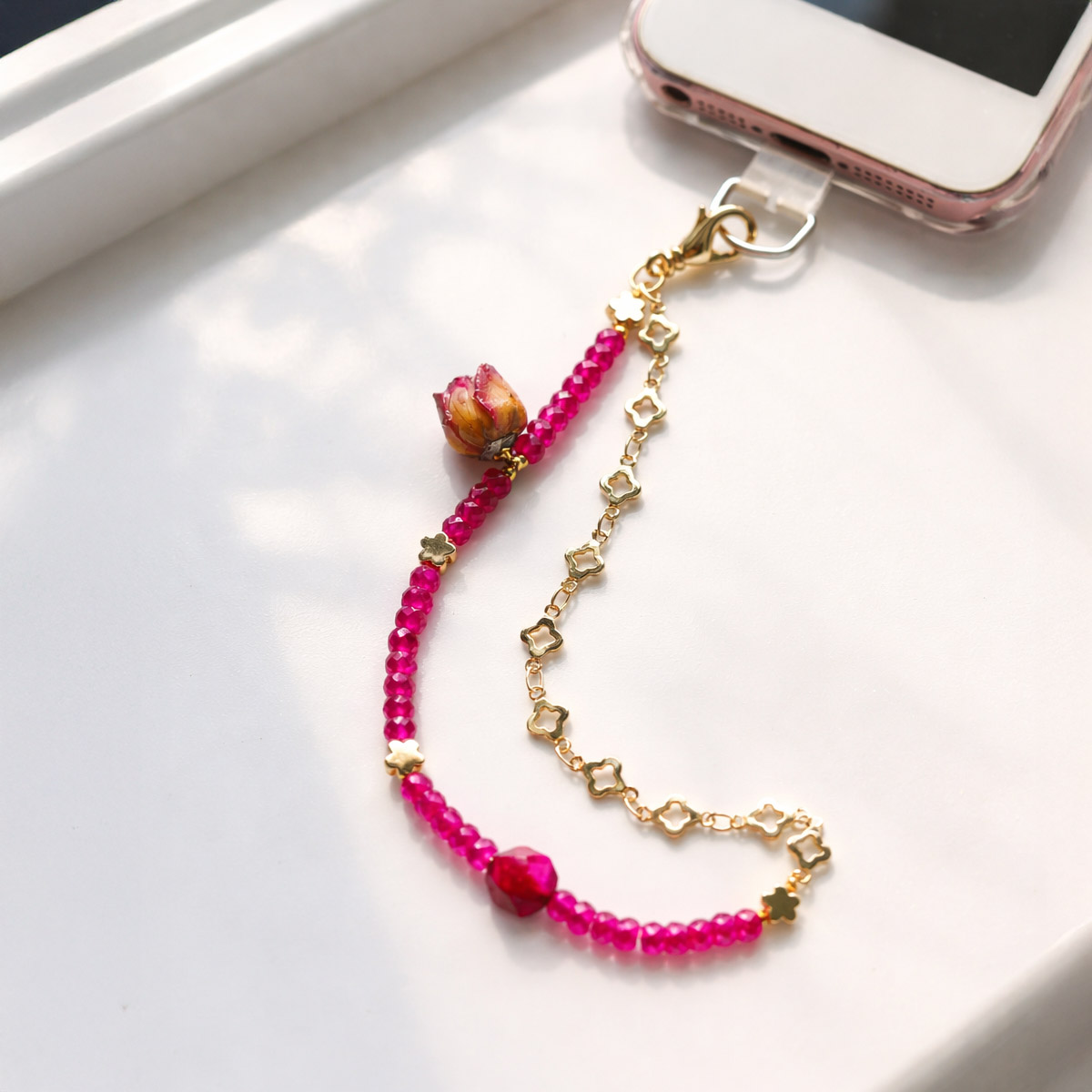 natu天然紅剛石玫瑰手機掛繩，紅剛石切面珠搭配乾燥玫瑰花與金色花形鍊條，窗邊自然光情境展示