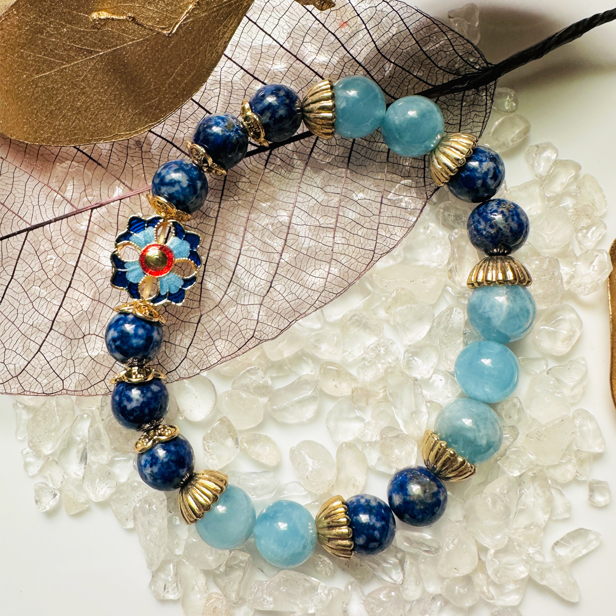 海藍寶景泰藍青金石手鍊情境擺拍,手鍊置於白水晶碎石與葉片之間,藍色系寶石搭配景泰藍花飾,呈現靜謐且具東方工藝感的風格