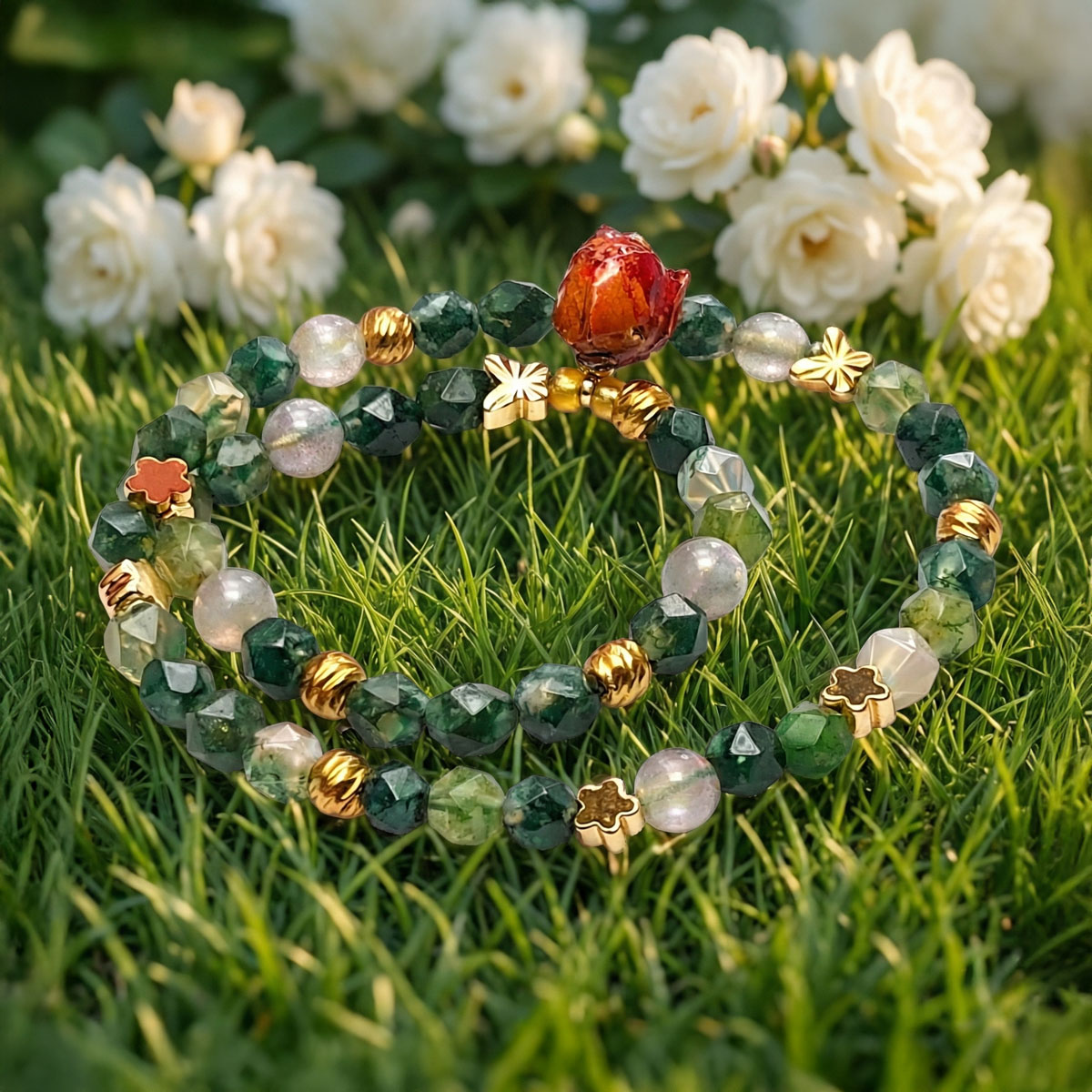 綠東菱玫瑰月光雙圈鍊置於翠綠草地與白玫瑰花叢中，綠色水晶與金色配件在陽光下呈現清新柔和的自然氛圍。