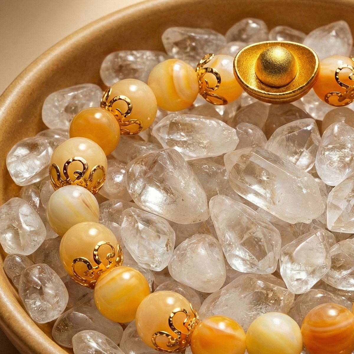 暖陽黃玉元寶手鍊置於盛滿透明水晶碎石的聚寶盆中,溫潤黃玉珠搭配金色元寶設計,整體色調柔和穩定,呈現富足與安心的視覺氛圍