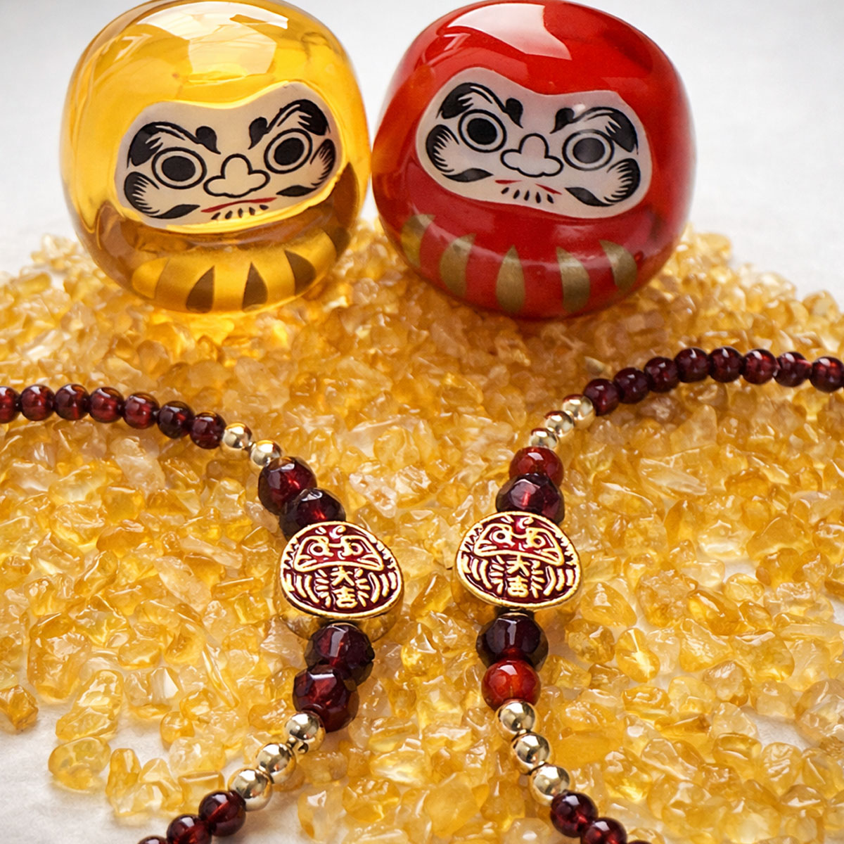 紅石榴石達摩不倒翁手鍊情境展示,搭配達摩擺件與黃色水晶背景,象徵穩定與祝福意象