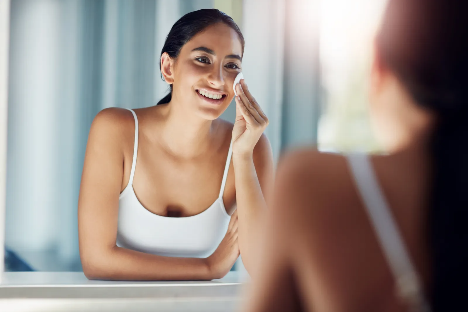 Shot of a beautiful young woman cleaning her face with cotton wool in the bathroom mirror